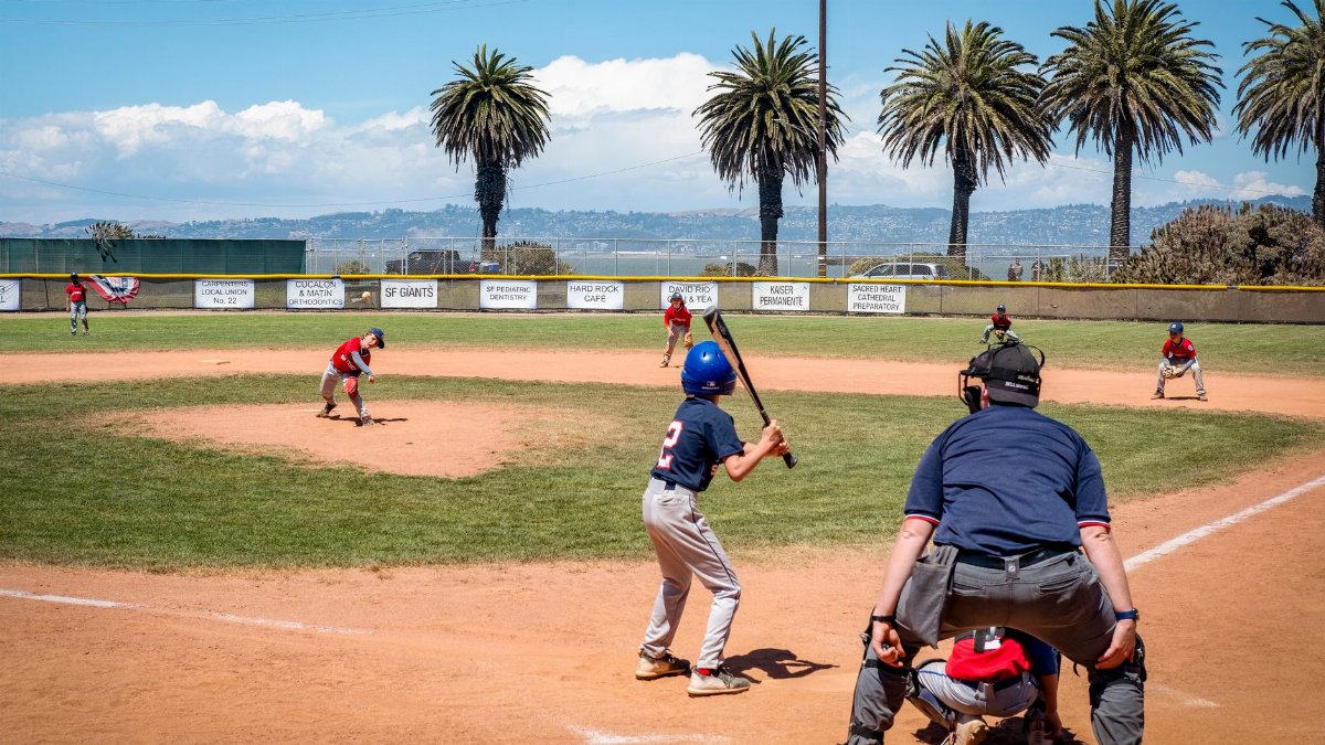 Children playing baseball in an outdoor field under a bright blue sky with palm trees in the background.