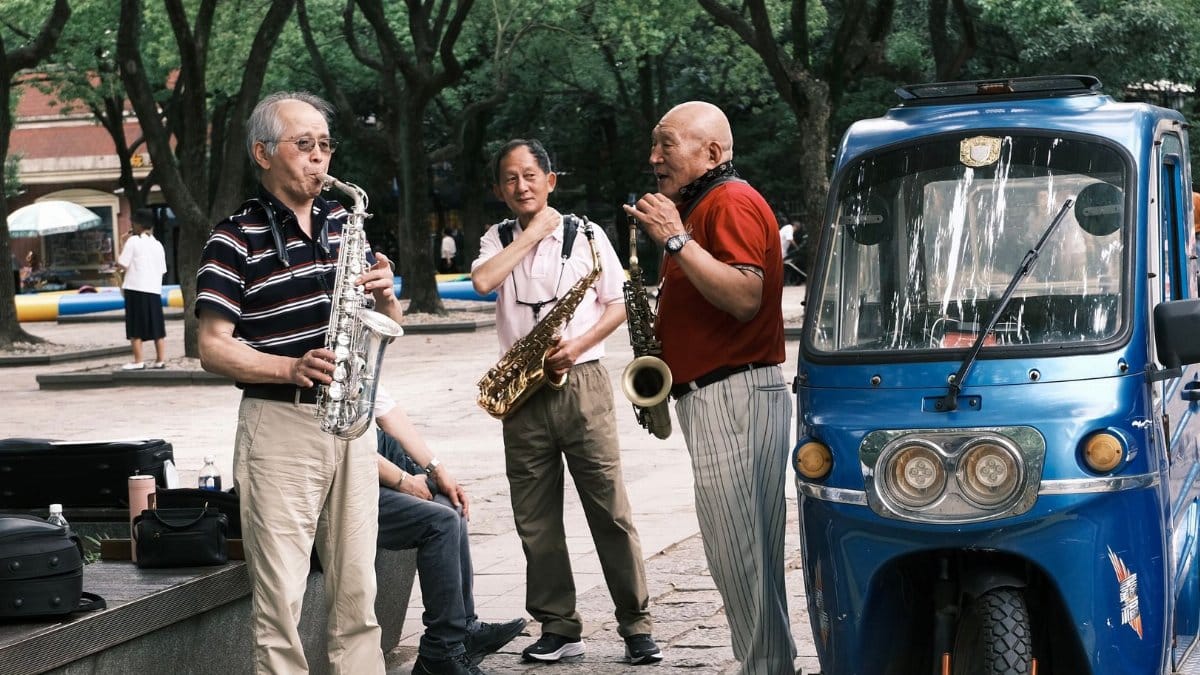 Asian men play saxophones on a city street, performing next to a blue vehicle.