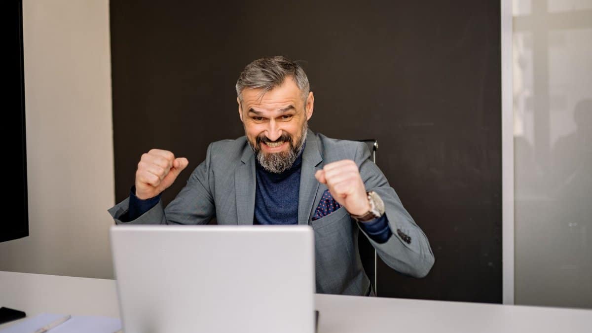 Happy businessman celebrating success while looking at laptop in modern office.