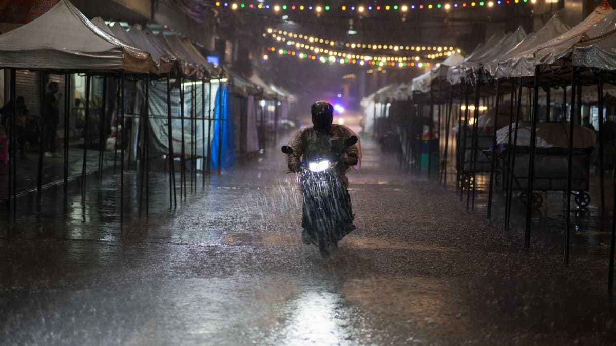 Motorcycle rider braves heavy rain in Bangkok street lined with tents and string lights at night.