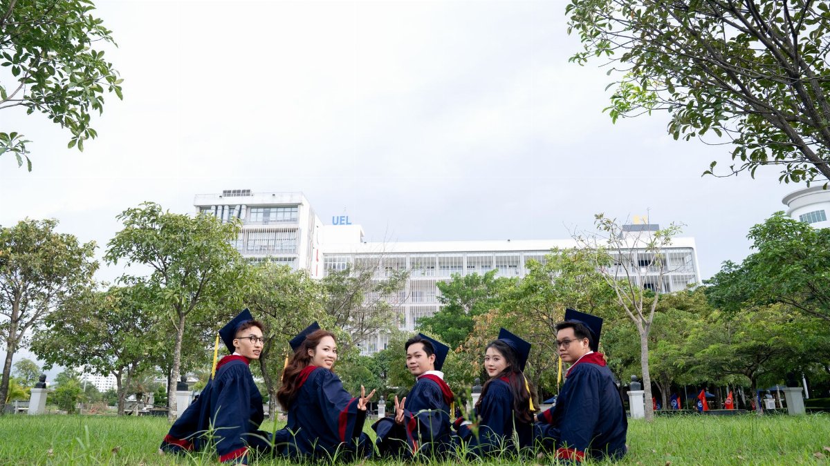 Group of five university graduates celebrating outdoors in regalia, sitting with a campus backdrop.