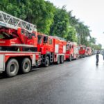 A long line of bright red fire trucks parked on a tree-lined street, indicating emergency preparedness.