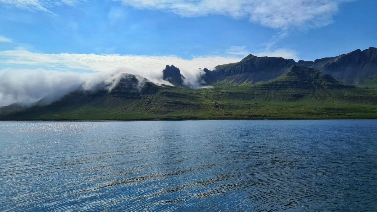 Picturesque view of rippling blue water surface with reflection of blue sky and shore with green hills slopes under clouds