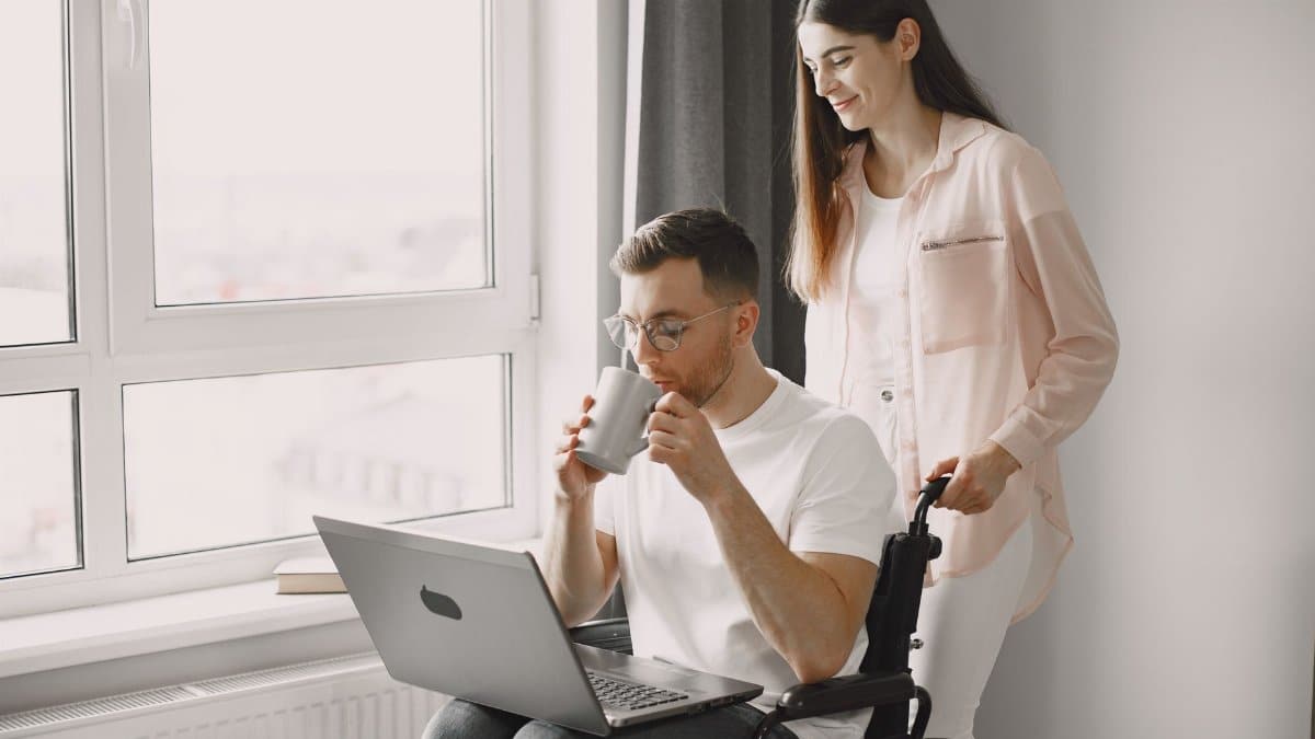 A caring woman assists a man in a wheelchair as they enjoy coffee and work on a laptop indoors.