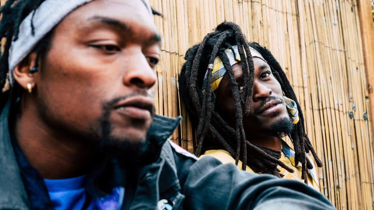 Two men with dreadlocks and headbands lean against a bamboo wall outdoors, captured in an urban setting.
