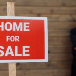 A confident real estate agent stands by a home for sale sign against a wooden wall.
