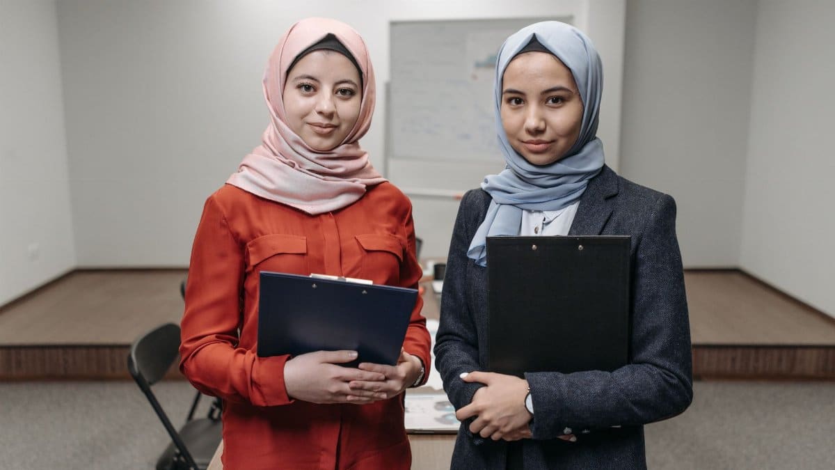 Two Muslim businesswomen in office attire with clipboards, indoors.