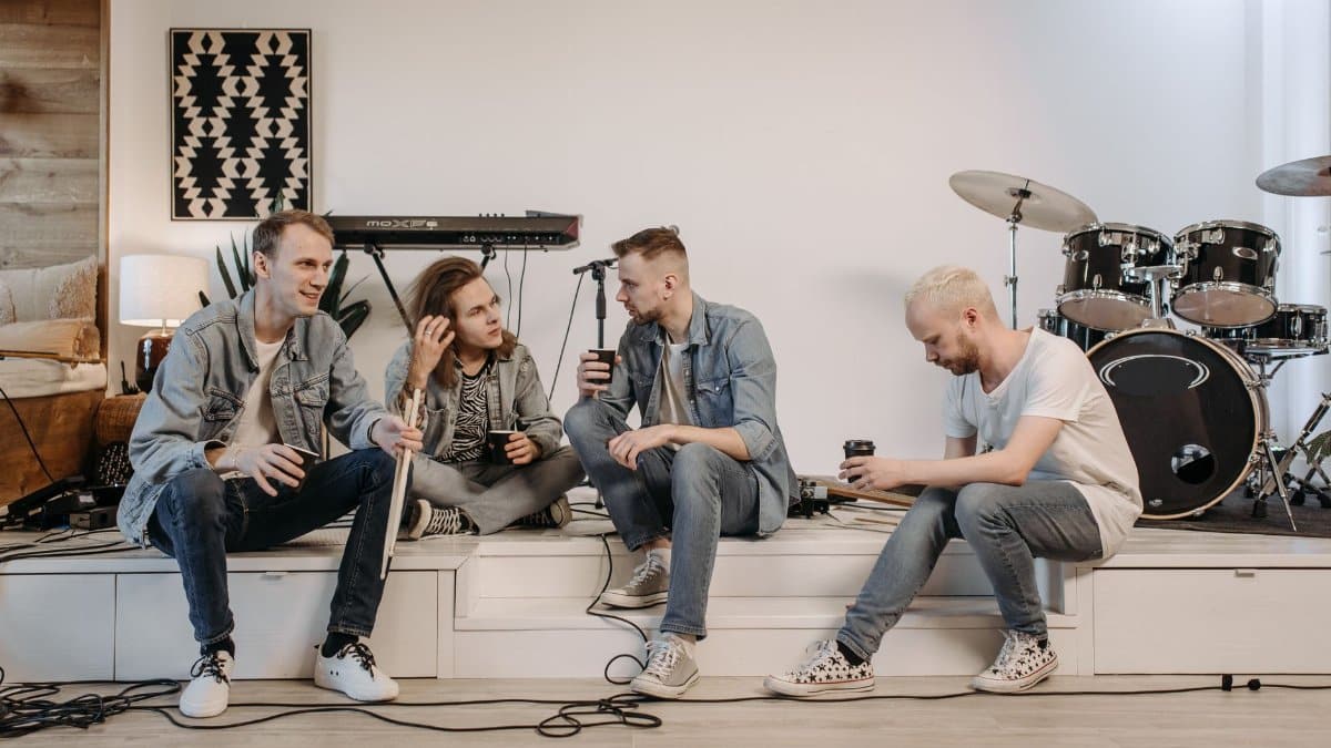 A band of four musicians sitting with coffee mugs, chatting in an indoor studio.