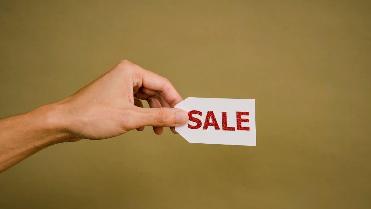 Crop anonymous person showing small white paper tag with inscription Sale against brown wall