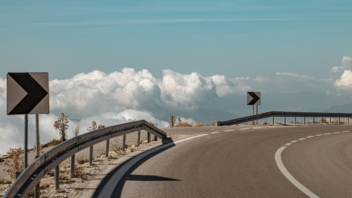 Curved mountain road with signposts, bordered by clouds and blue sky.