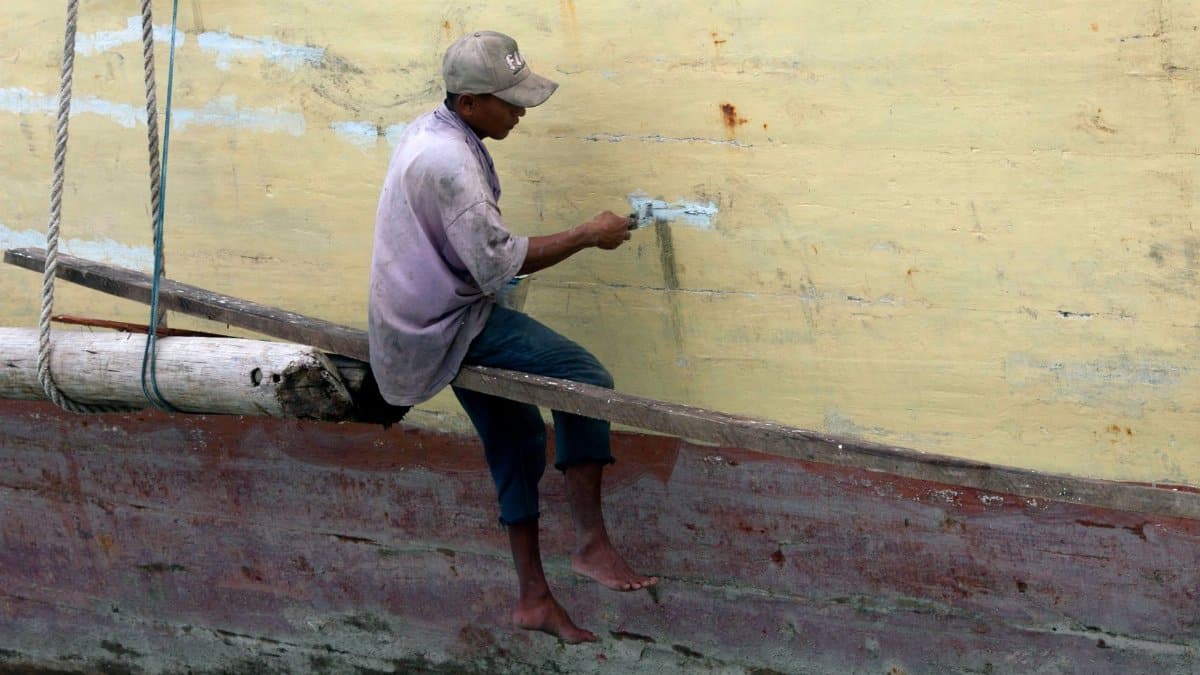 A man is painting a boat hull, balancing on a plank outdoors.