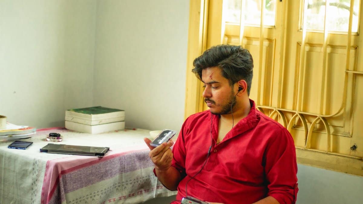 A young man in red shirt listening to a tape recorder by a window in a sunlit room.