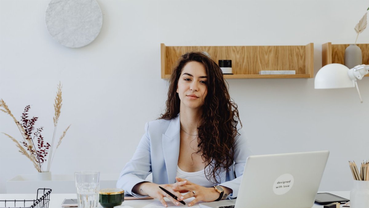 Businesswoman in a light blazer sitting at a desk with a laptop, exuding confidence in a modern office.