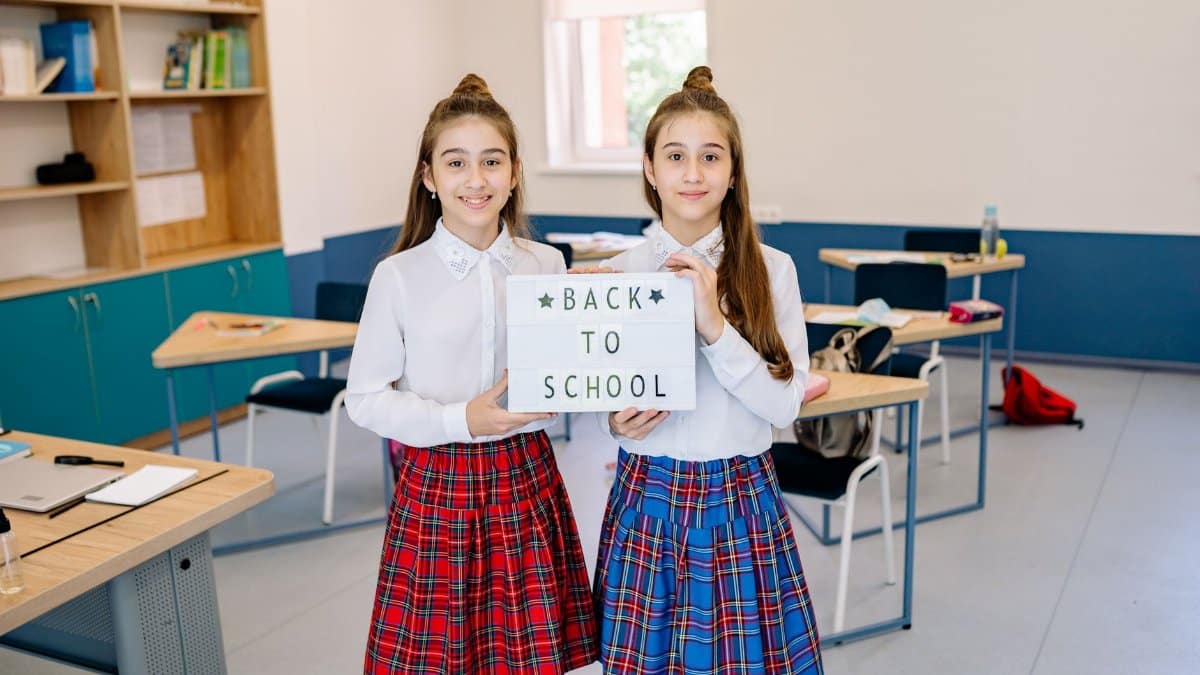 Twin sisters in uniforms holding a back to school sign, standing in a classroom.