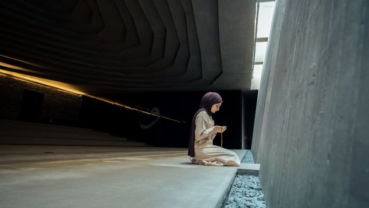 A young Middle Eastern woman in a hijab praying indoors, holding prayer beads.