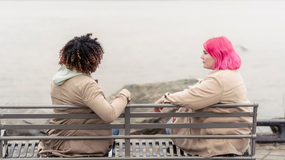 Two women wearing coats chat on a bench by a river, reflecting leisure and friendship.