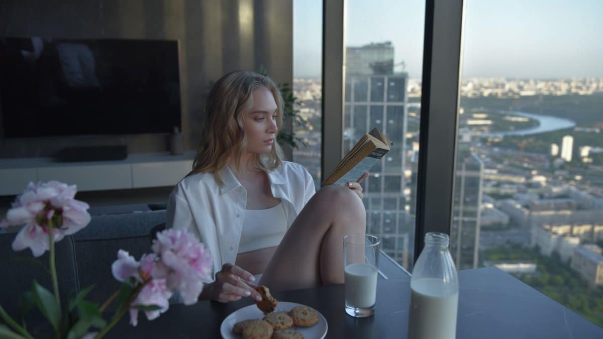 Relaxing scene of a woman reading by a city view, enjoying cookies and milk.