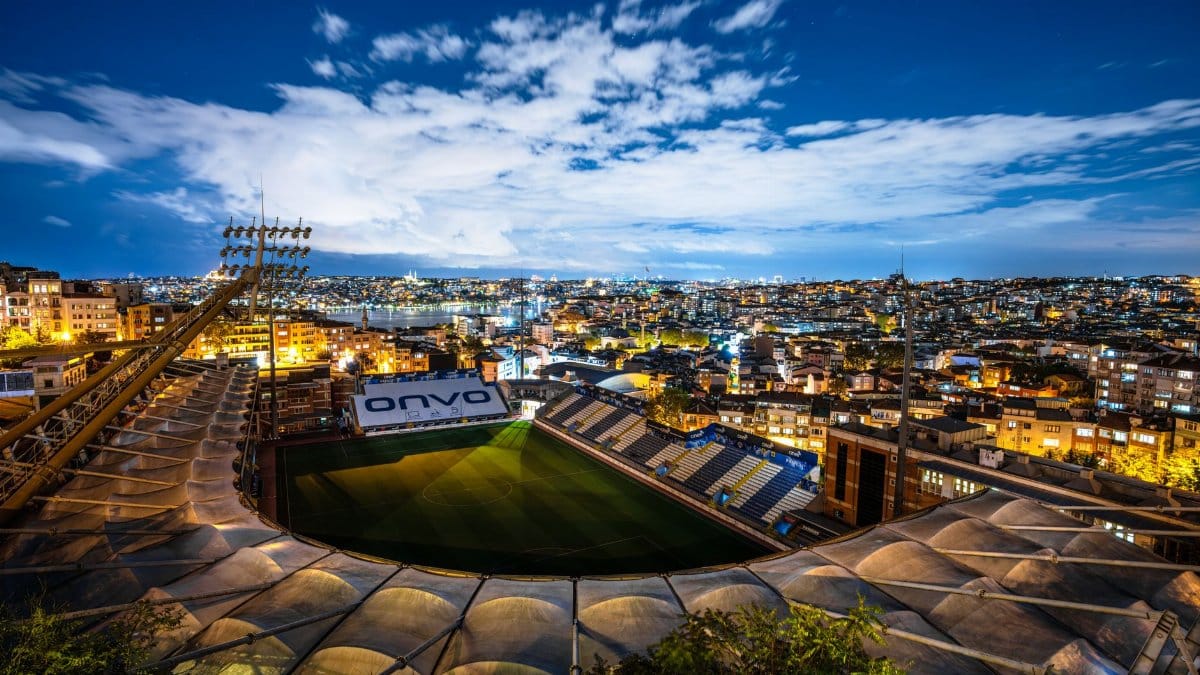 Aerial view of a brightly lit stadium surrounded by city buildings at dusk under a clear sky.