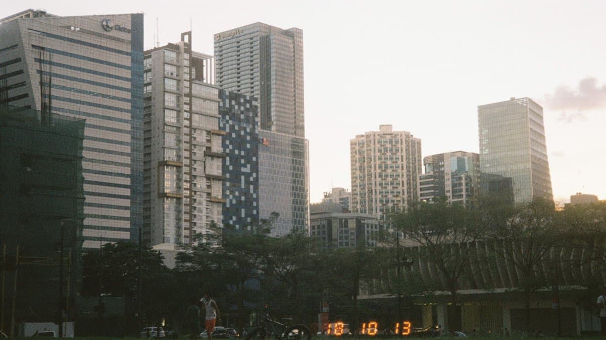 City skyline at sunset with modern skyscrapers and greenery in the foreground.