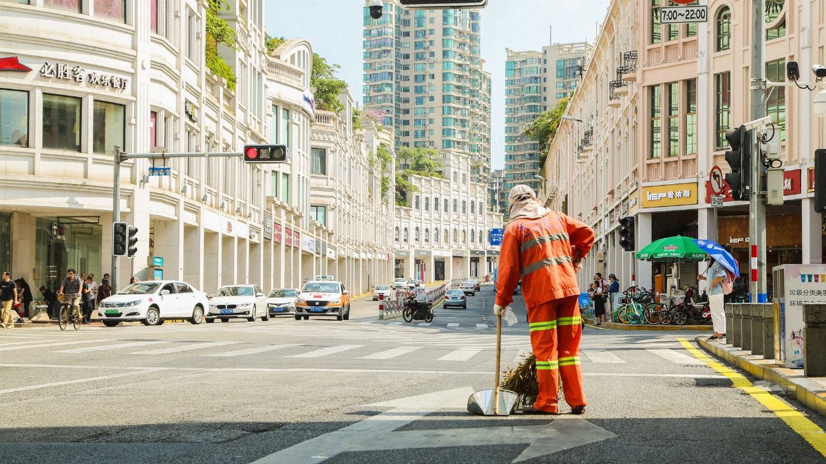 A city street cleaner in orange uniform at a busy intersection with cars and buildings.