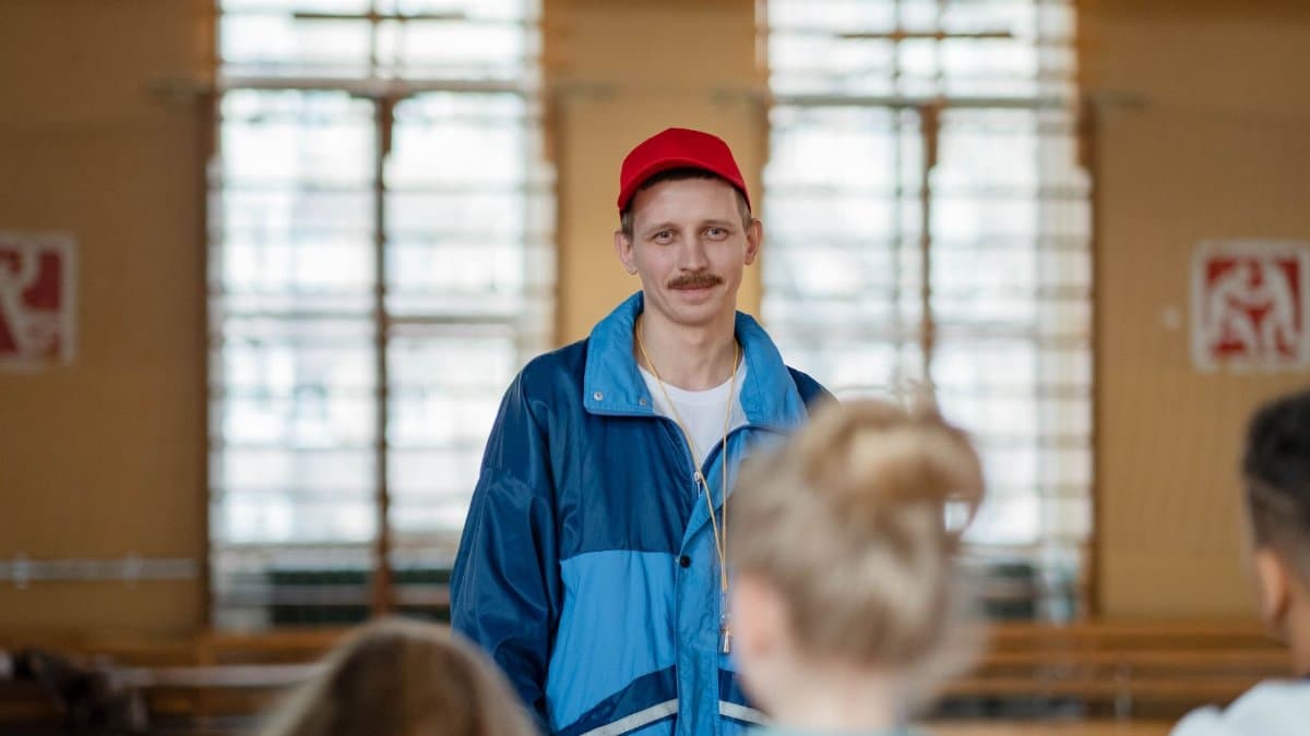 Male coach in a gym engaging with a group of children, wearing casual sportswear.