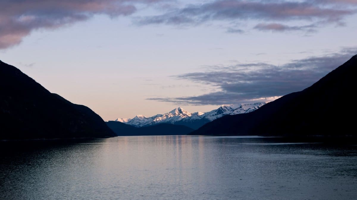 Tranquil view of snowcapped mountains reflecting in a serene lake under a dawn sky in Skagway, Alaska.