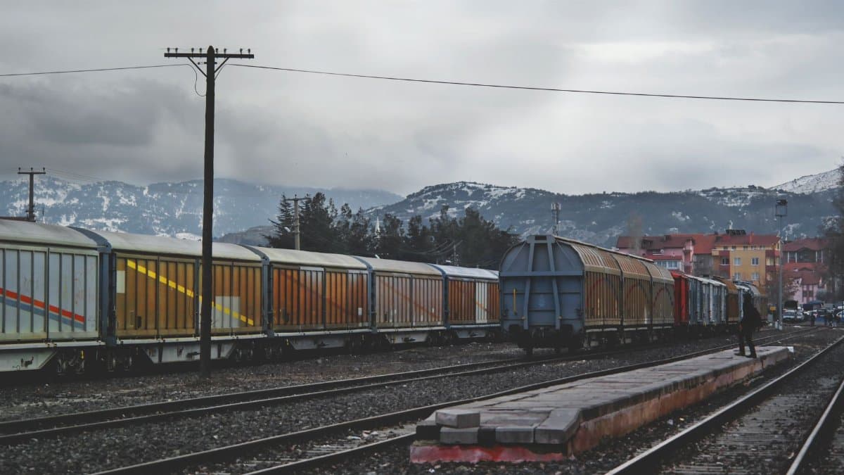A freight train with colorful cargo wagons on a mountain railway track under a gray sky.