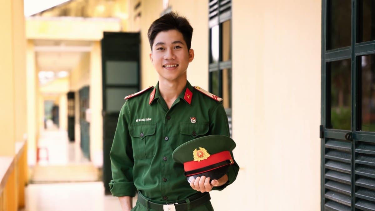 A young soldier in uniform smiling while standing outdoors, holding a cap.