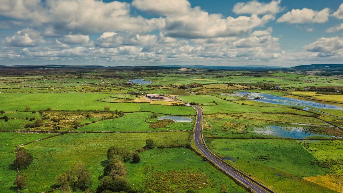 Aerial view of the vibrant green countryside and long road in County Clare, Ireland, under a partly cloudy sky.