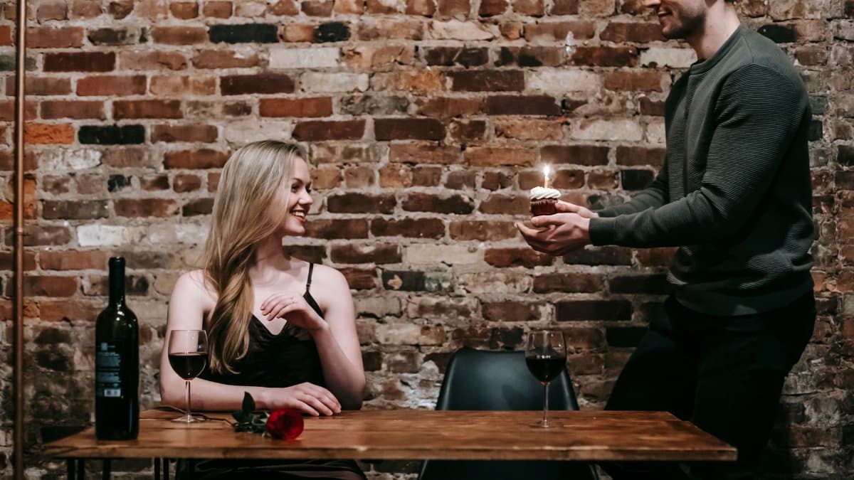 Happy young man giving woman birthday cupcake and candle on date in restaurant at table with glasses and bottle with red wine and rose near brick wall