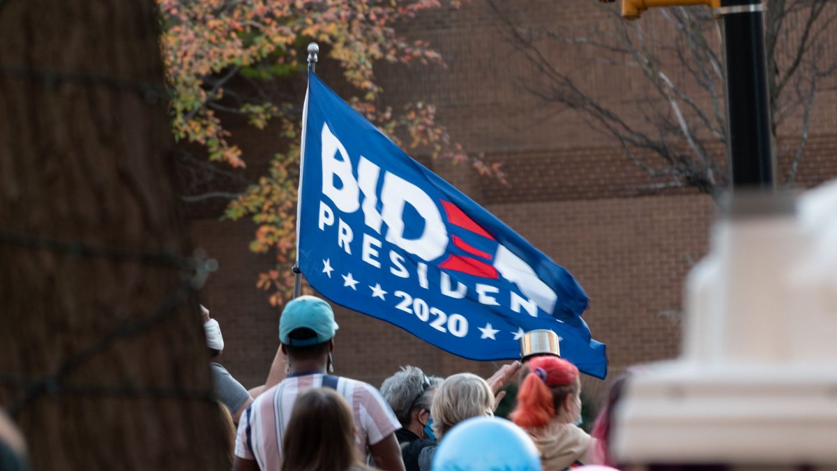 A diverse crowd gathered outdoors holding a Biden 2020 flag during a protest.