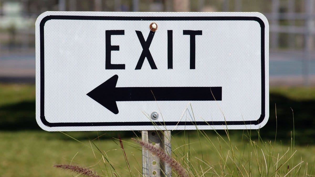 Exit sign with left-pointing arrow in a grassy outdoor setting.