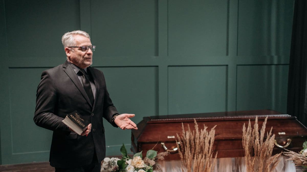 A solemn funeral ceremony featuring a pastor in a black suit delivering a speech next to a wooden coffin adorned with flowers.