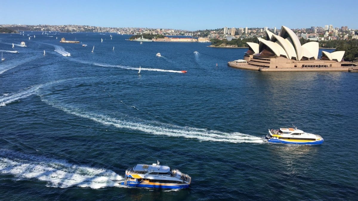 Aerial view of Sydney Harbour featuring the Opera House and ferries under a clear blue sky.