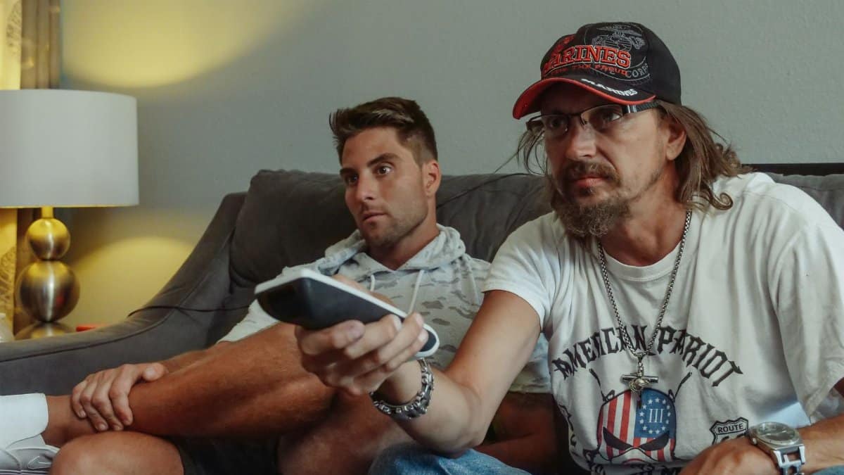 Two men relaxing on a sofa at home, watching TV with a remote control in hand.