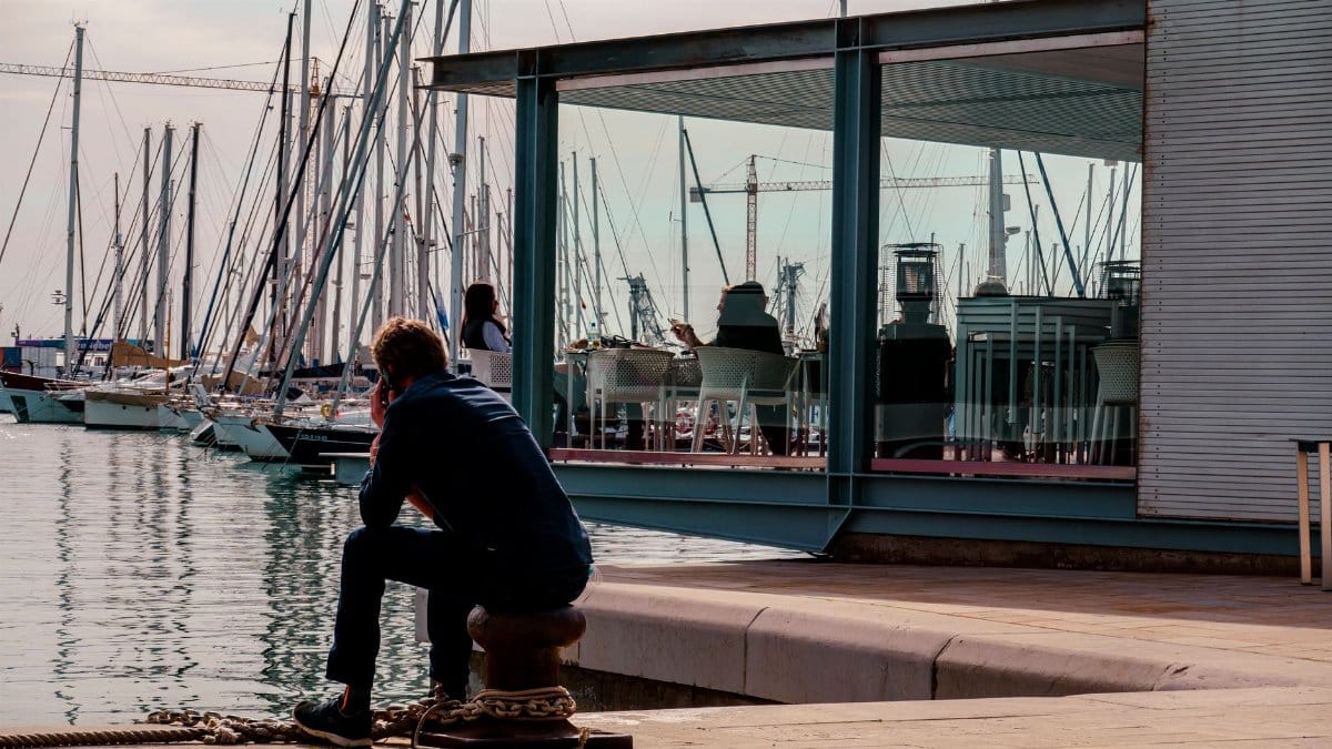 Man seated by Alicante harbor, enjoying the summer view of moored boats and reflections.