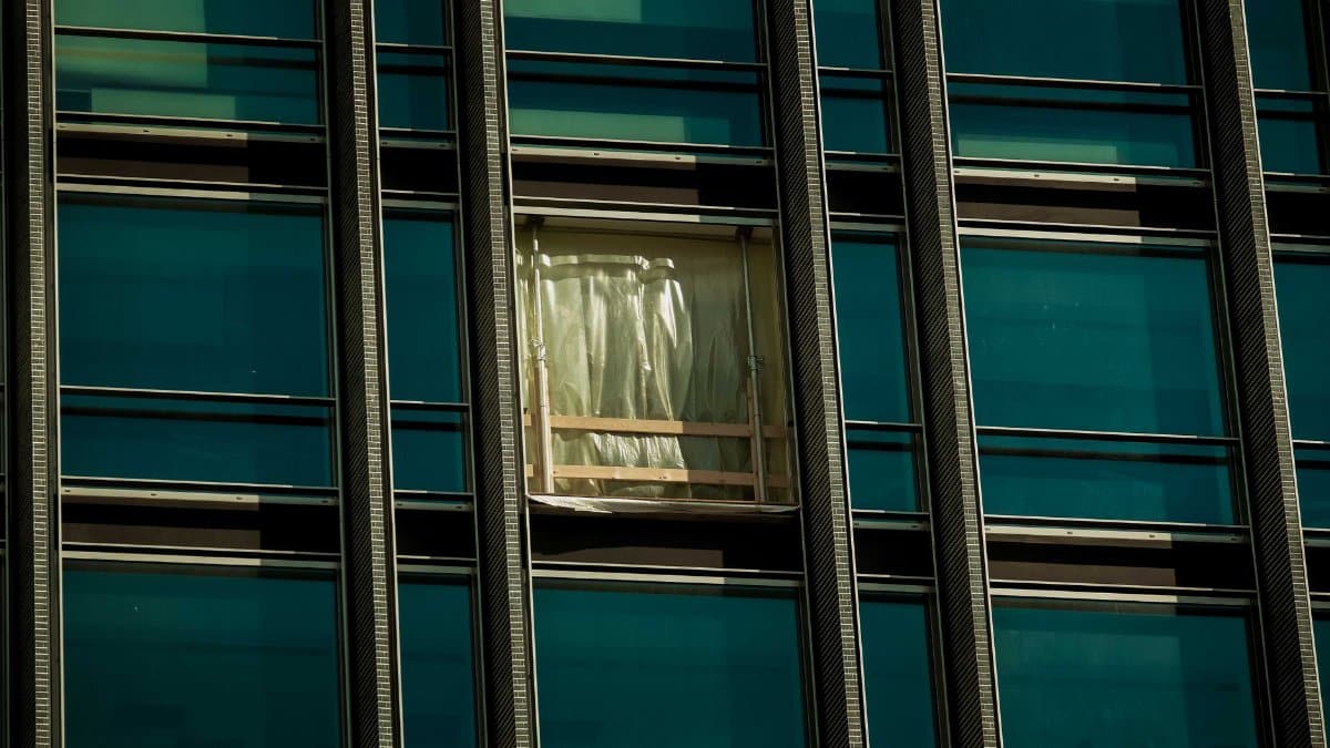 A close-up view of a modern skyscraper's facade with a focus on a broken window covered with a temporary plastic sheet.