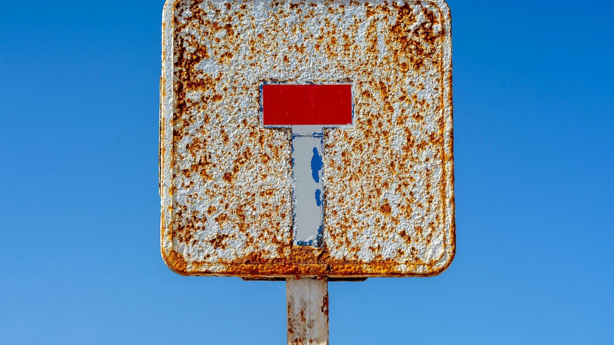 A rusty dead-end sign against a clear blue sky symbolizing caution and neglect.