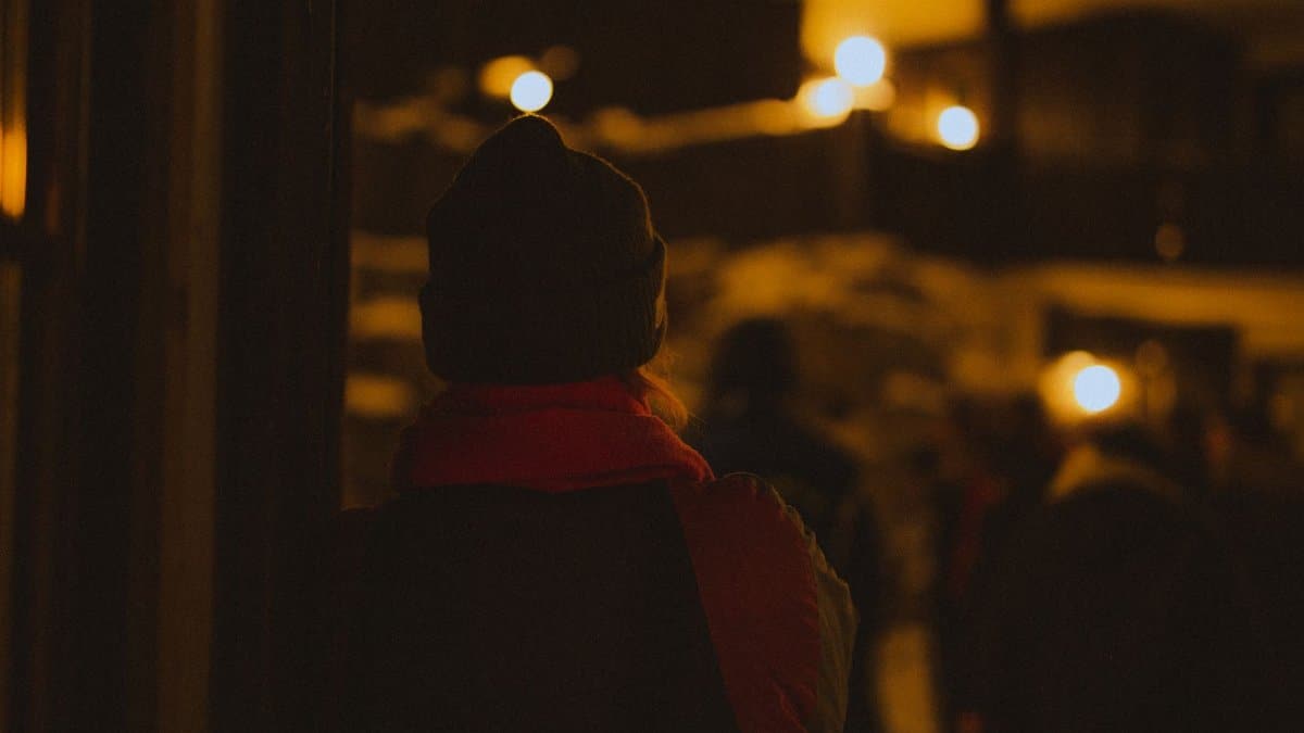 A person wearing a beanie and scarf is silhouetted against warm outdoor night lights.