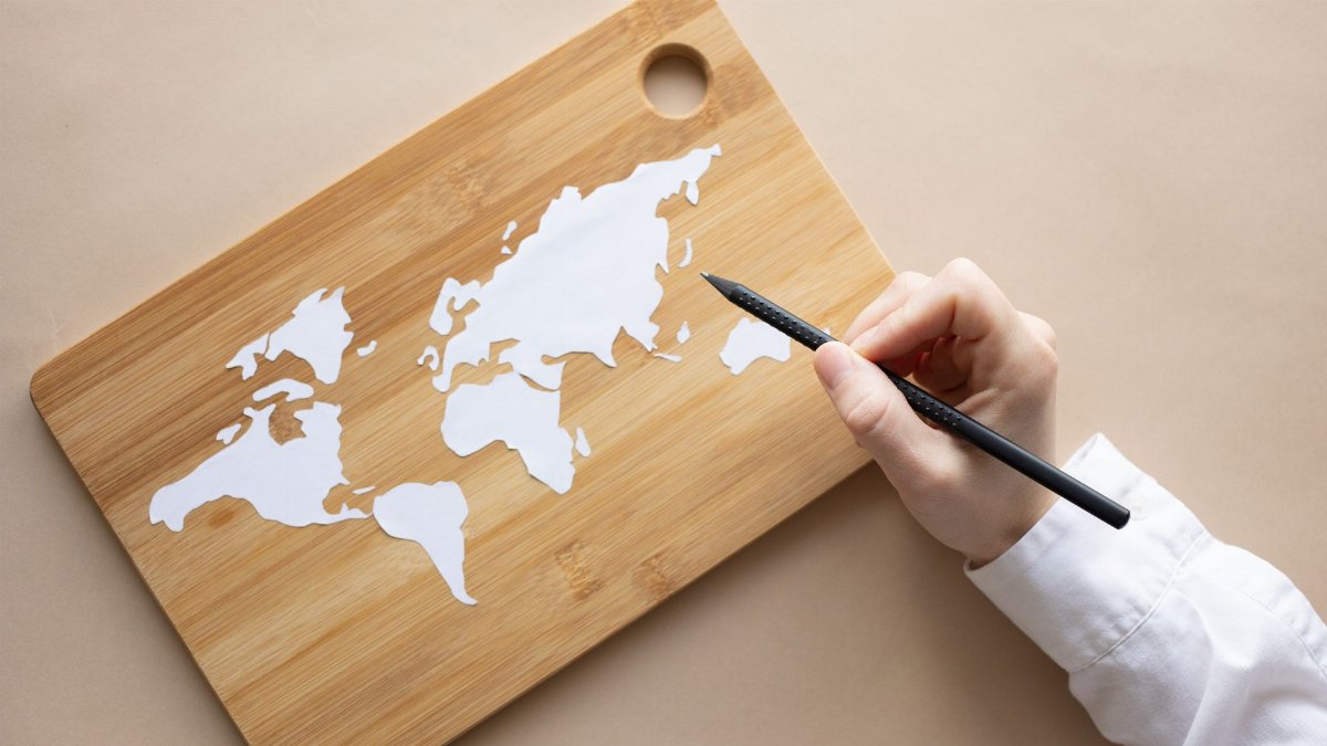 Top view of crop anonymous artist drawing world map on wooden desk with white pencil