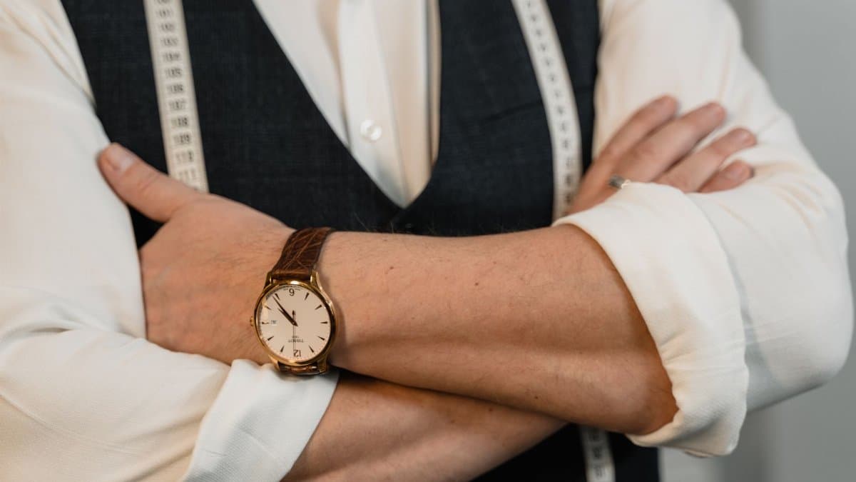 A tailor crosses arms showing a stylish wristwatch and tape measure, representing precision craftsmanship.