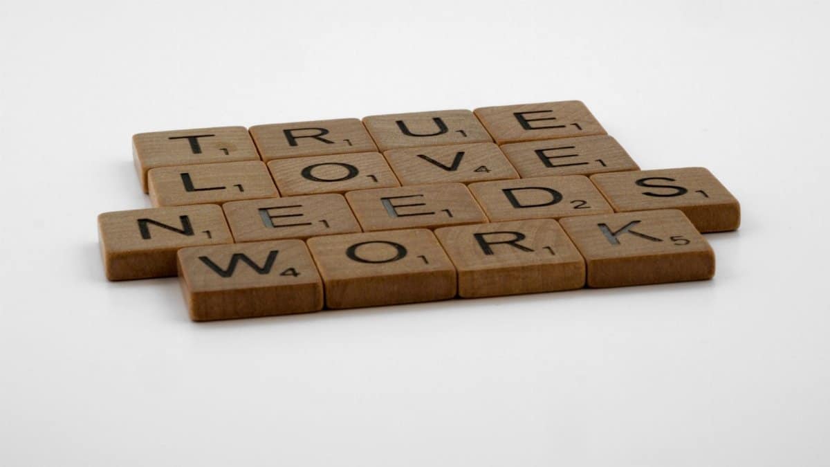 Wooden Scrabble tiles on white background spelling 'True Love Needs Work.'