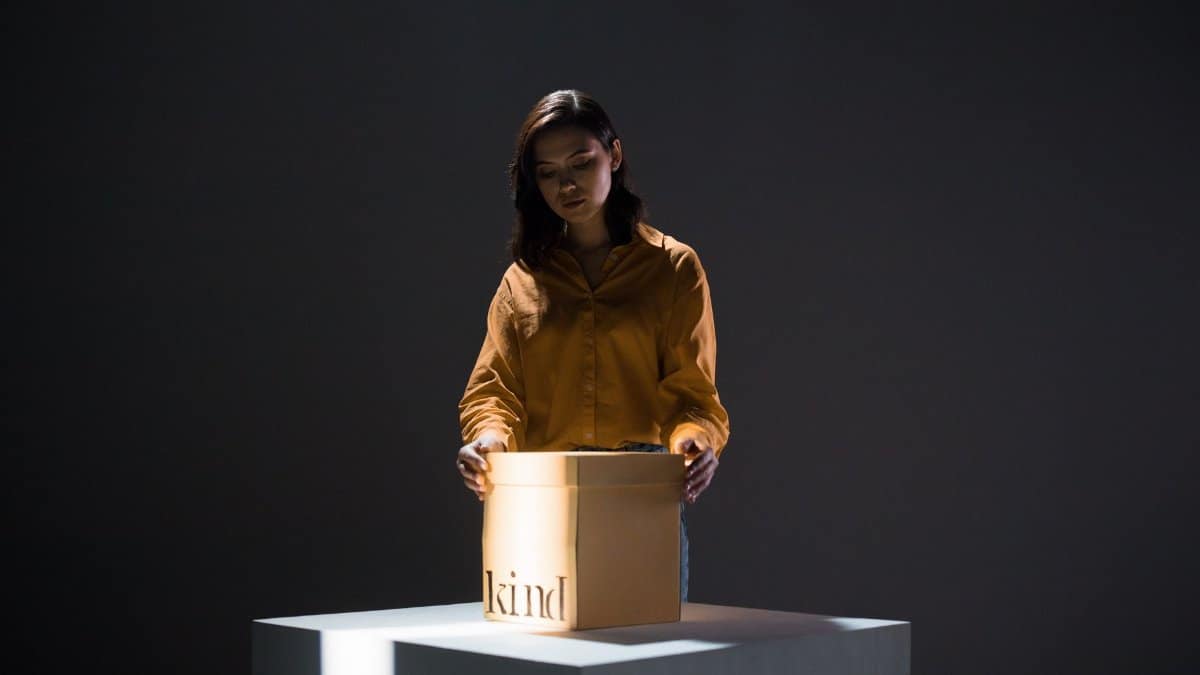 A woman stands before an illuminated box labeled 'kind' in a dimly lit studio, symbolizing hope and charity.
