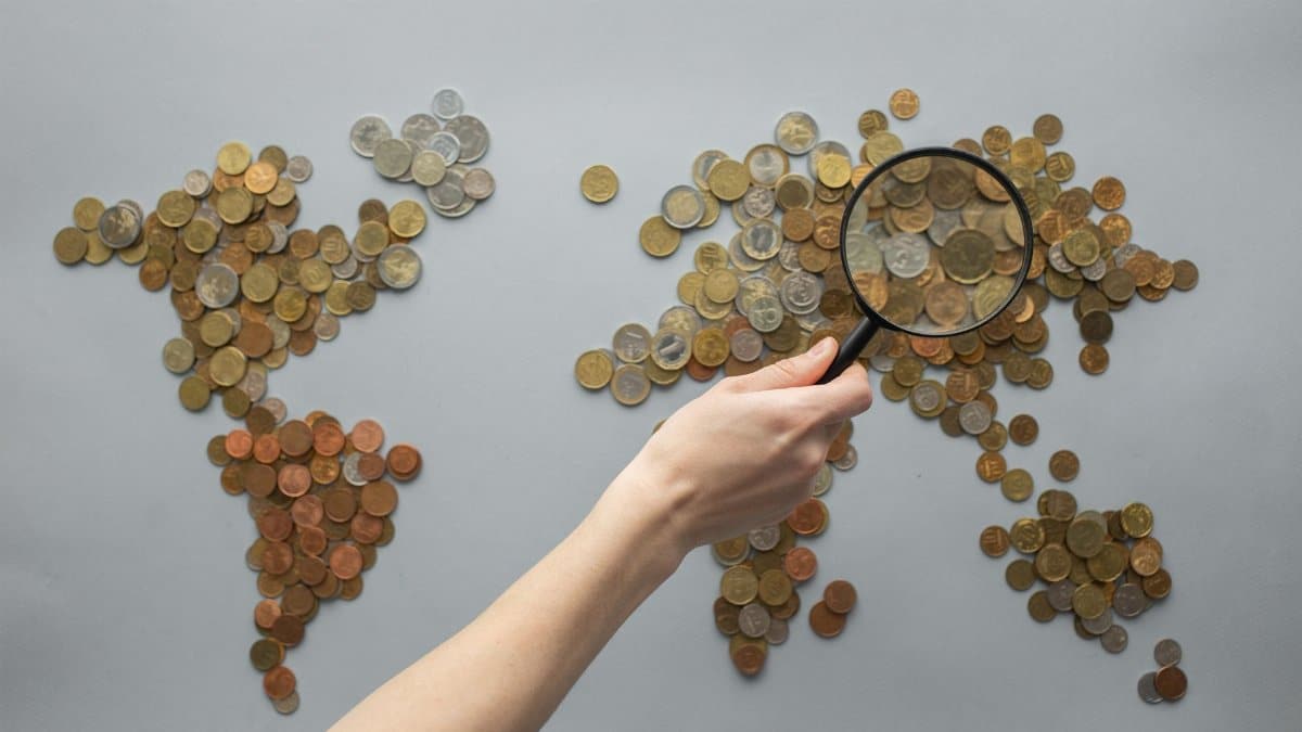 Top view of crop unrecognizable traveler with magnifying glass standing over world map made of various coins on gray background