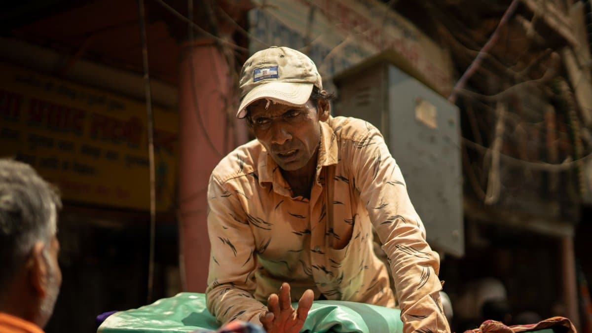 A street vendor wearing a cap sells colorful textiles in a bustling urban market setting.