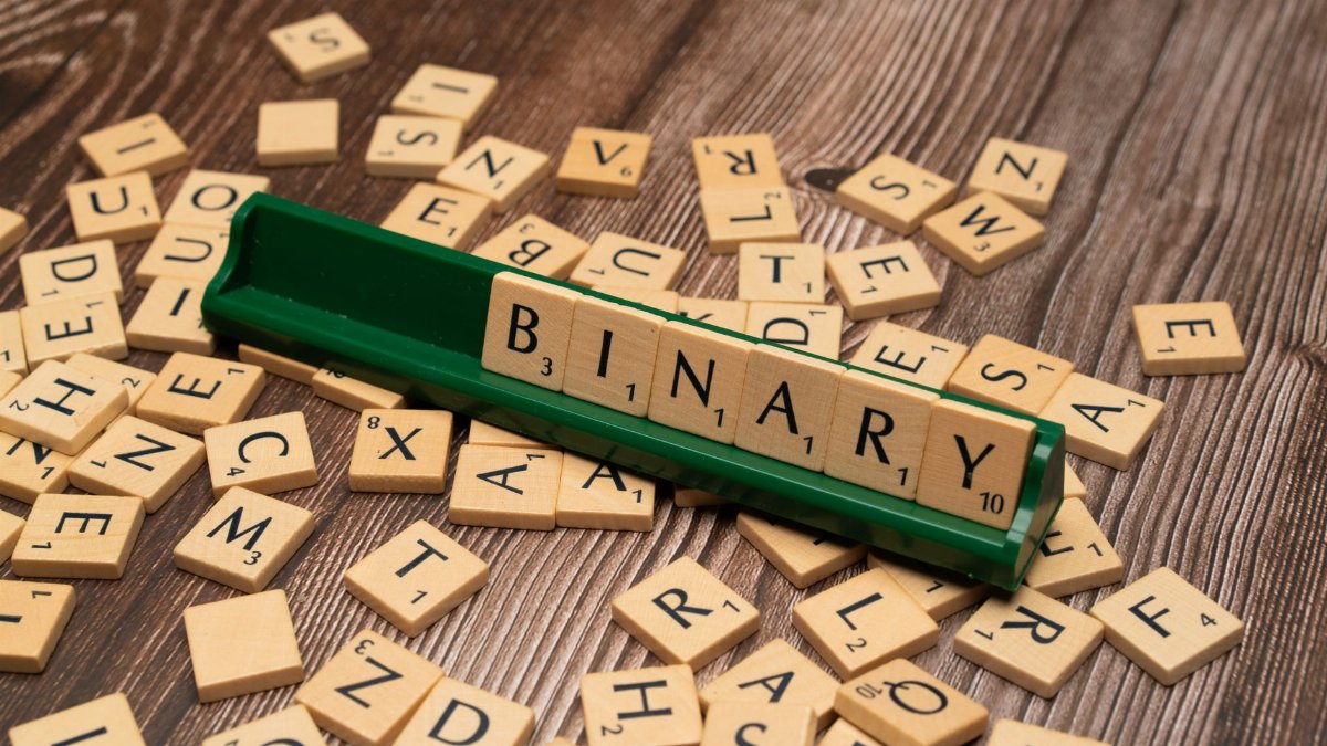 Close-up of scrabble tiles spelling 'Binary' surrounded by scattered letters on a wooden background.