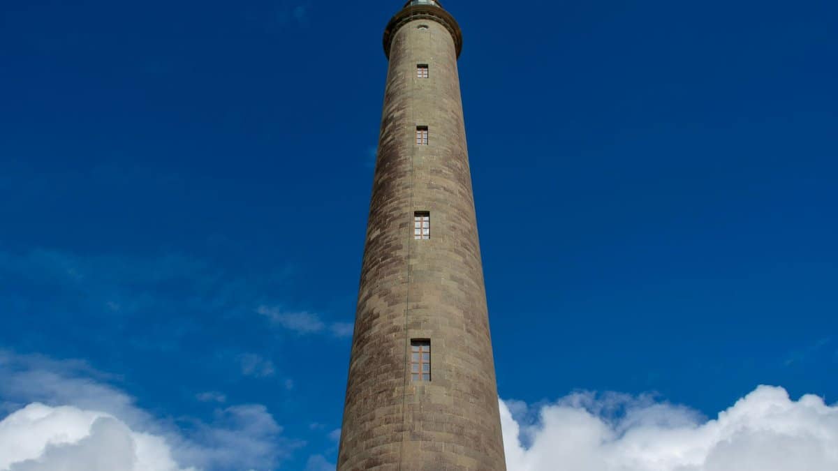 Dramatic low-angle shot of Maspalomas Lighthouse against a clear blue sky with fluffy clouds.