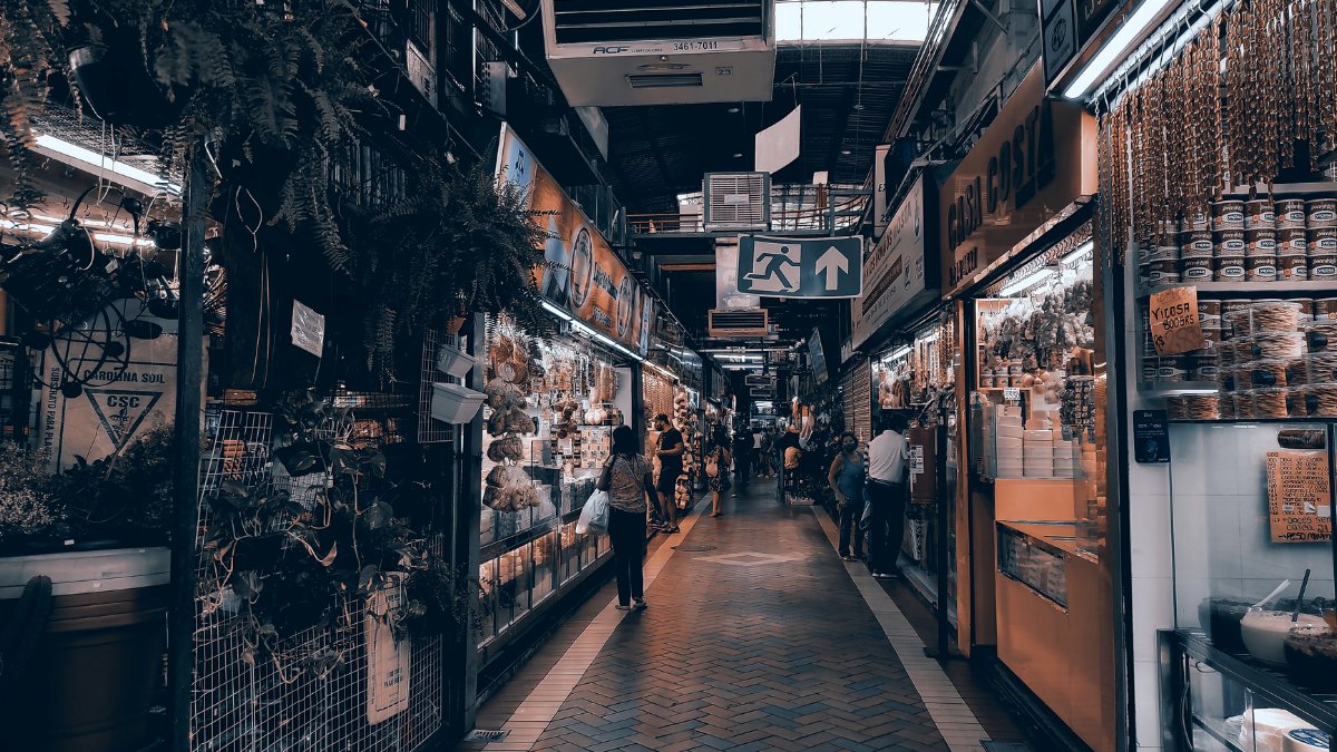 A vibrant indoor market scene with various stalls and shoppers exploring the goods on a busy pathway.