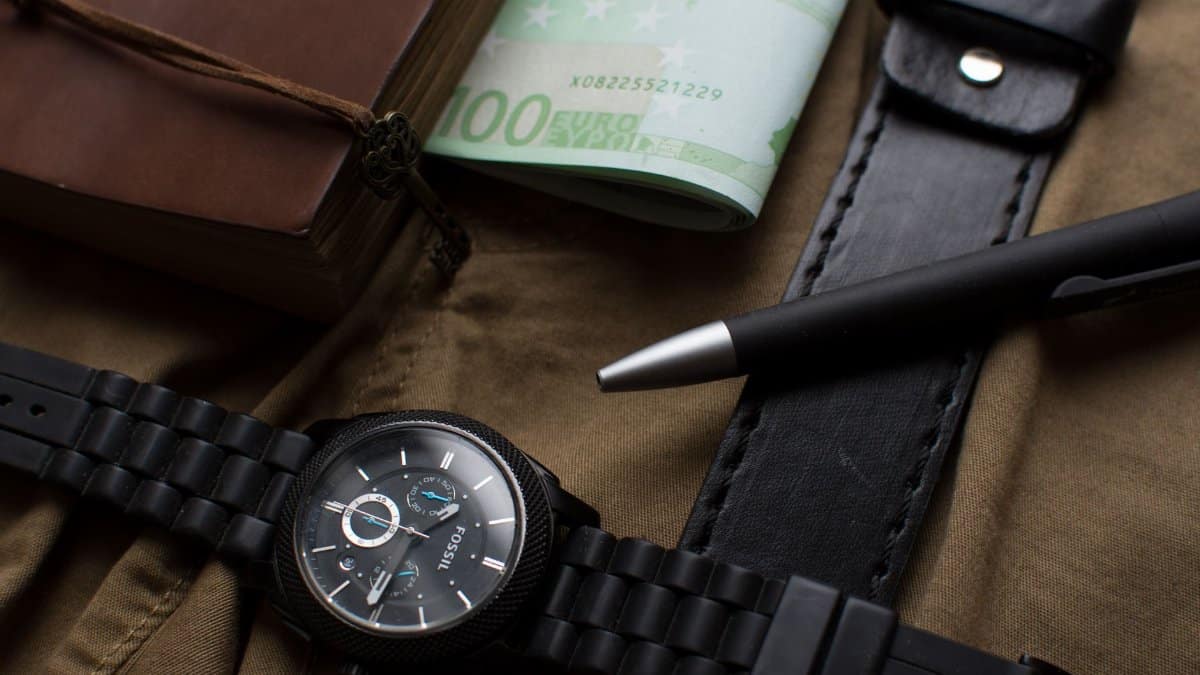 Flat lay featuring a watch, euros, leather belt, and a pen on a dark background.