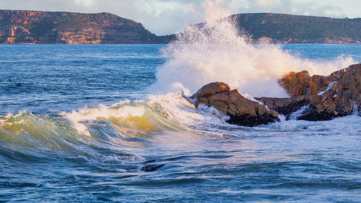Powerful ocean waves crash against rocks at Plettenberg Bay, South Africa, under a bright sky.